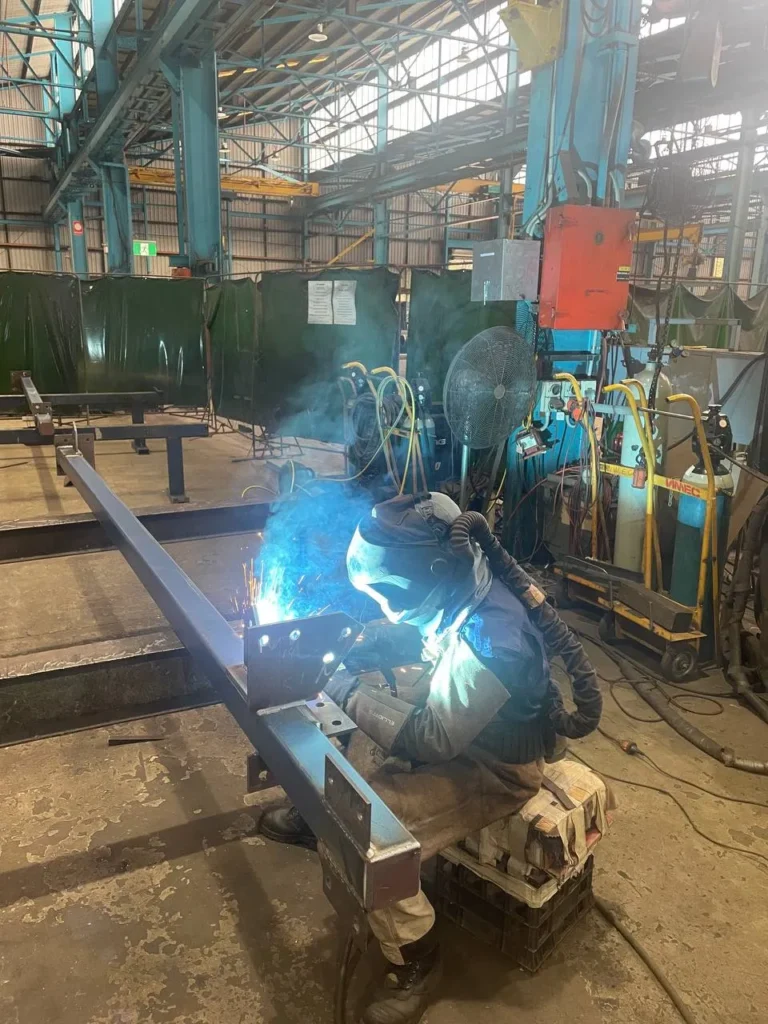 Welder in a helmet welding a prototype steel beam while the weld is witnessed