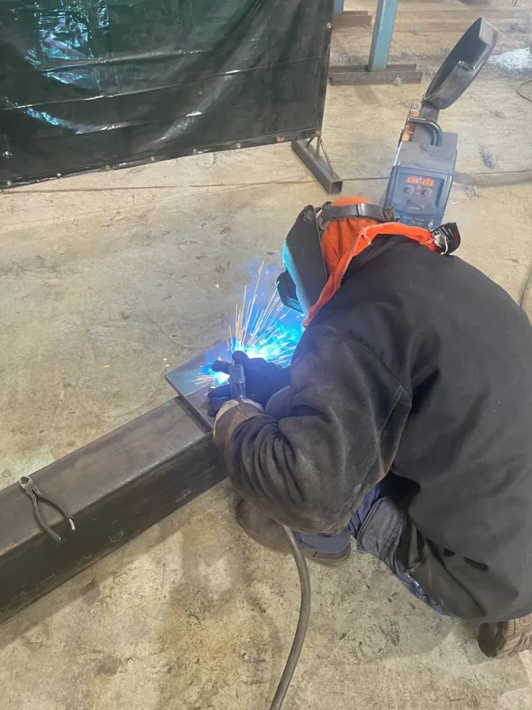 Welder in a helmet and protective jacket welding a steel section under supervision